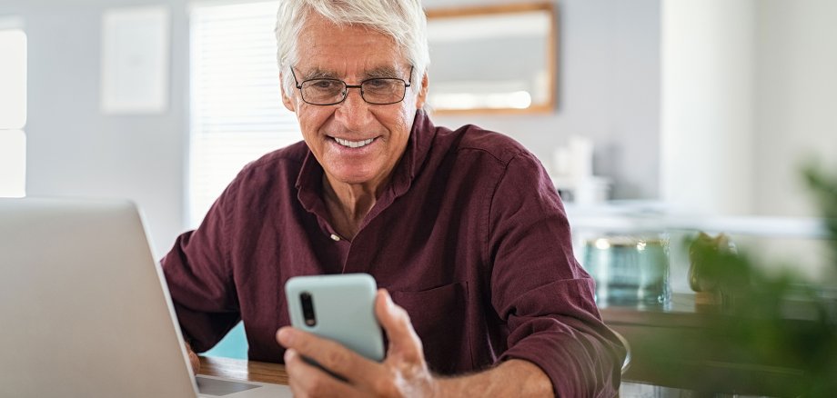  Seniorer Mann, der an einem Laptop arbeitet und ein Smartphone benutzt.