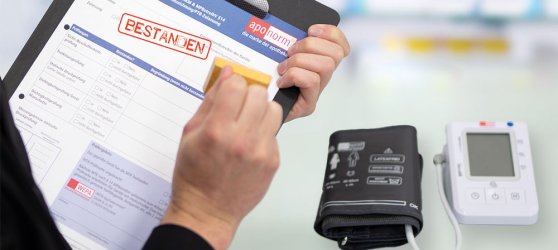 FAQ Checkup Eine Frau mit einem Klemmbrett hält gerade einen Stempel in der Hand, um ein Blatt zu stempeln wo mit rot "Bestanden" steht. Im Vordergrund ist ein Blutdruckmessgerät zu sehen, und im Hintergrund erkennt man unscharf eine Apotheke