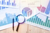 Adobestock Viele Blätter mit Diagrammen und Anzeichen, die auf einem Tisch liegen, und eine Lupe sowie ein Stift liegen auf den Blättern.