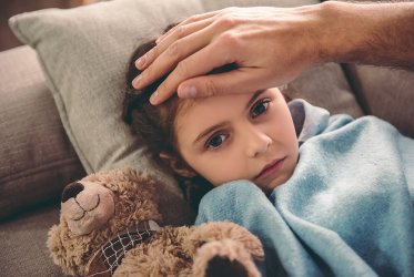 Fieber Ein Kind, das auf dem Sofa liegt, in einer blauen Decke, mit einer Hand an ihrem Kopf, misst Fieber. Sie sieht krank aus.