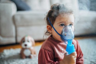Inhalator Ein kleines Kind, das eine Inhalationsmaske trägt und diese festhält, aus der Dampf kommt. Im Hintergrund ist ein Abriss eines Wohnzimmers zu sehen.