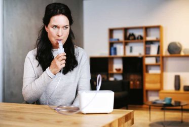 Inhalator Eine Frau mit schwarzen Haaren sitzt an einem Tisch und inhalisiert mit einem Mundstück aus dem Inhalationsgerät, das auf dem hölzernen Tisch steht. Im Hintergrund ist ein Abriss eines Wohnzimmers zu erkennen.